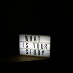 A glowing light box sign asks "what is your story"