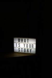 A glowing light box sign asks "what is your story"